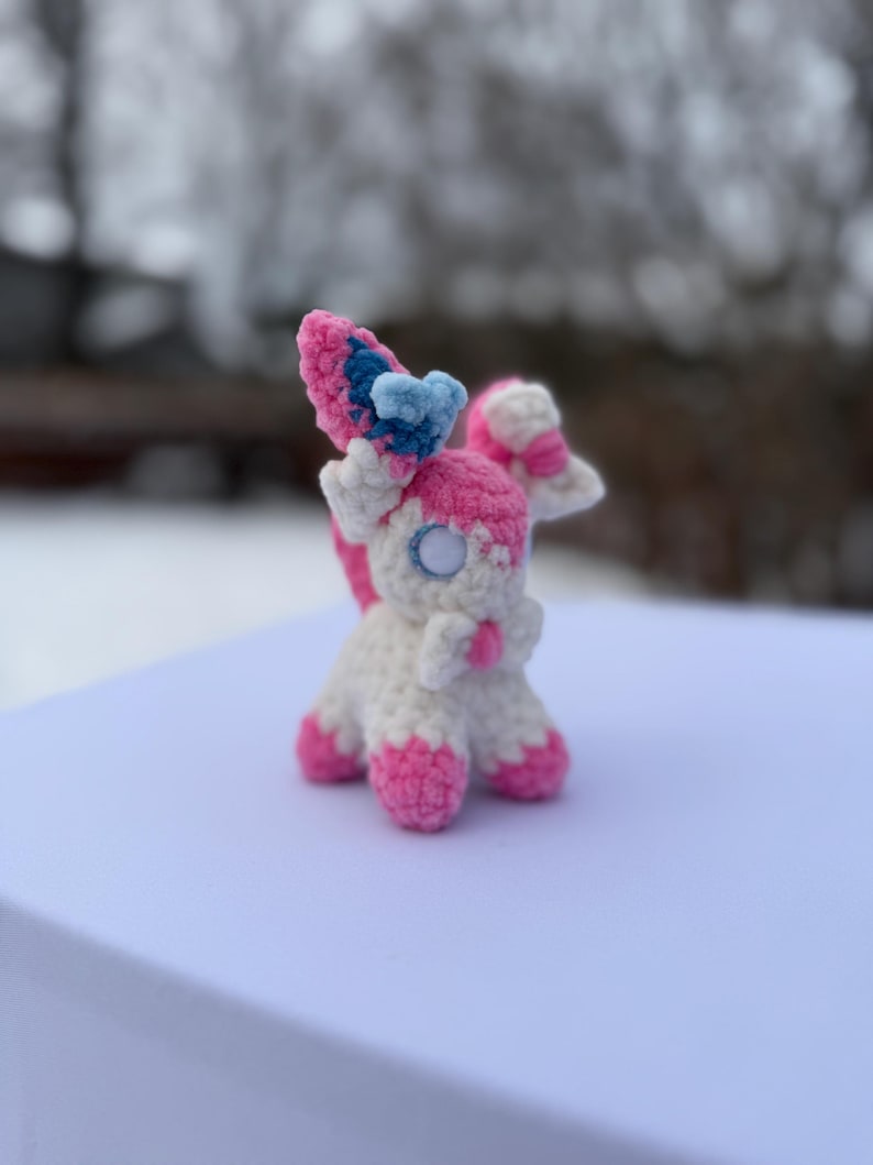 Puede incluir: Un peluche Sylveon hecho a mano con ganchillo. El peluche presenta un cuerpo blanco con detalles rosas en los pies, las orejas y la cola. Las orejas son rosas y azules, y los ojos son blancos con un centro negro. El peluche est&aacute; sobre una superficie blanca.