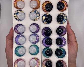 May include: A sheet of white cardstock with 24 round, colorful, resin-filled, glitter-covered, moon-shaped, eye-like embellishments. The embellishments are arranged in four rows of six. Each row features a different color and glitter combination.