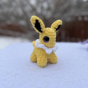 Pu&ograve; includere: Un giocattolo di peluche all'uncinetto giallo fatto a mano con grandi orecchie, dettagli neri e occhi viola. Ha un colletto a balze bianco ed &egrave; appoggiato su una superficie innevata. Il giocattolo &egrave; una rappresentazione di un personaggio popolare.