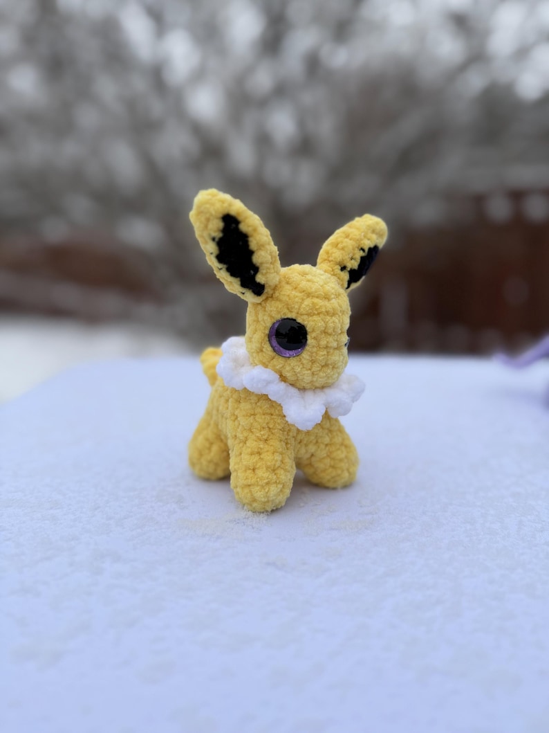 Pu&ograve; includere: Un giocattolo di peluche all'uncinetto giallo fatto a mano con grandi orecchie, dettagli neri e occhi viola. Ha un colletto a balze bianco ed &egrave; appoggiato su una superficie innevata. Il giocattolo &egrave; una rappresentazione di un personaggio popolare.