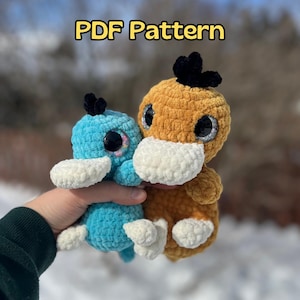 May include: Two crocheted plush toys in the shape of platypuses. One is blue with white accents and the other is brown with white accents. Both have large black eyes. The image includes the text "PDF Pattern".