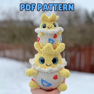 Puede incluir: Un juguete de peluche tejido a crochet de un personaje amarillo de Pokemon, con una cáscara de huevo blanca y detalles azules y rojos. El juguete está sentado en una mano, con una versión más pequeña del mismo juguete encima. La imagen incluye el texto "PDF PATTERN".
