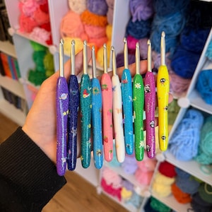 May include: A collection of ten metal crochet hooks with colourful, glittery, and patterned handles. The handles feature various designs including planets, mermaids, strawberries, butterflies, flowers, and pandas in shades of purple, blue, pink, white, green, and yellow.