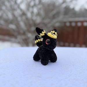 Pu&ograve; includere: Una piccola creatura all'uncinetto nera con dettagli gialli e neri sulle orecchie e sulla testa. Ha occhi rossi ed &egrave; posizionata su una superficie innevata. Il peluche fatto a mano &egrave; una creazione unica e stravagante.
