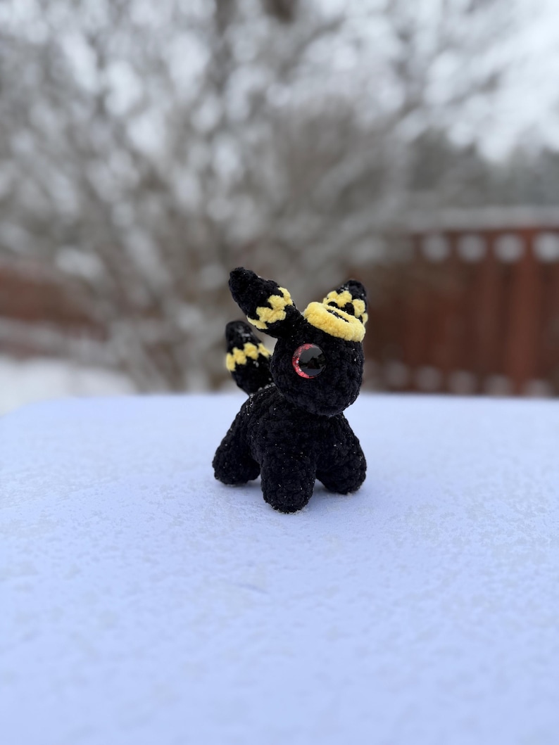 Pu&ograve; includere: Una piccola creatura all'uncinetto nera con dettagli gialli e neri sulle orecchie e sulla testa. Ha occhi rossi ed &egrave; posizionata su una superficie innevata. Il peluche fatto a mano &egrave; una creazione unica e stravagante.
