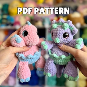 May include: Two handmade crochet dinosaur plushies in pink and purple. The pink dinosaur has blue accents, while the purple one has green accents. The text "PDF PATTERN" is displayed in the image.
