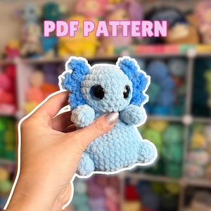 May include: A light blue crocheted axolotl plushie with dark blue frills and large black eyes. The text "PDF PATTERN" is displayed in pink above the plushie. The plushie is held in a hand.