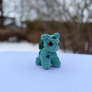 Może przedstawiać: Małe, turkusowe, szydełkowe stworzenie z ciemnoniebieskimi plamkami i r&oacute;żowymi oczami. Ma zieloną bulwę na plecach i siedzi na białej powierzchni. Tło jest rozmyte, z drzewami i zaśnieżonym krajobrazem.