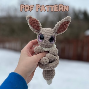 May include: A handmade, plush crochet bunny in shades of brown and beige, with large, glittery eyes. The bunny has long ears with a darker brown interior and a small, fluffy tail. The text "PDF PATTERN" is displayed at the top.