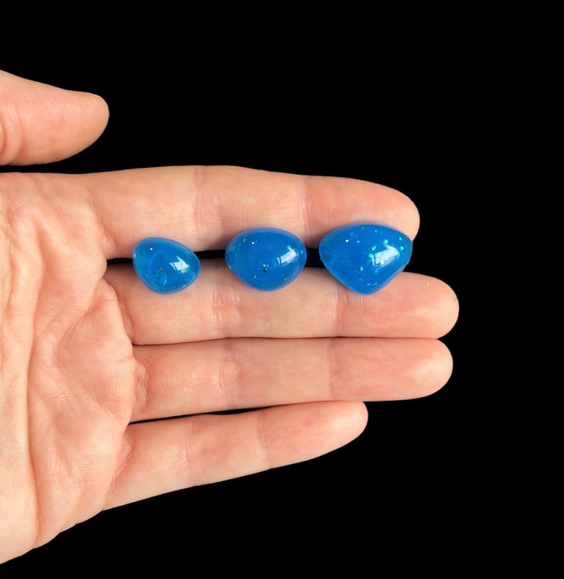 May include: Three blue resin cabochons of different sizes with a glittery finish. The cabochons are arranged in a row on a hand.