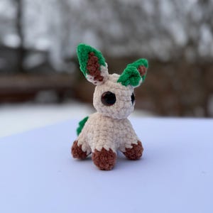Puede incluir: Un peluche hecho a mano con ganchillo en forma de un peque&ntilde;o animal. El juguete tiene un cuerpo color crema, patas marrones y orejas verdes y marrones. Tiene ojos negros grandes y una peque&ntilde;a cola verde. El juguete est&aacute; sobre una superficie blanca.