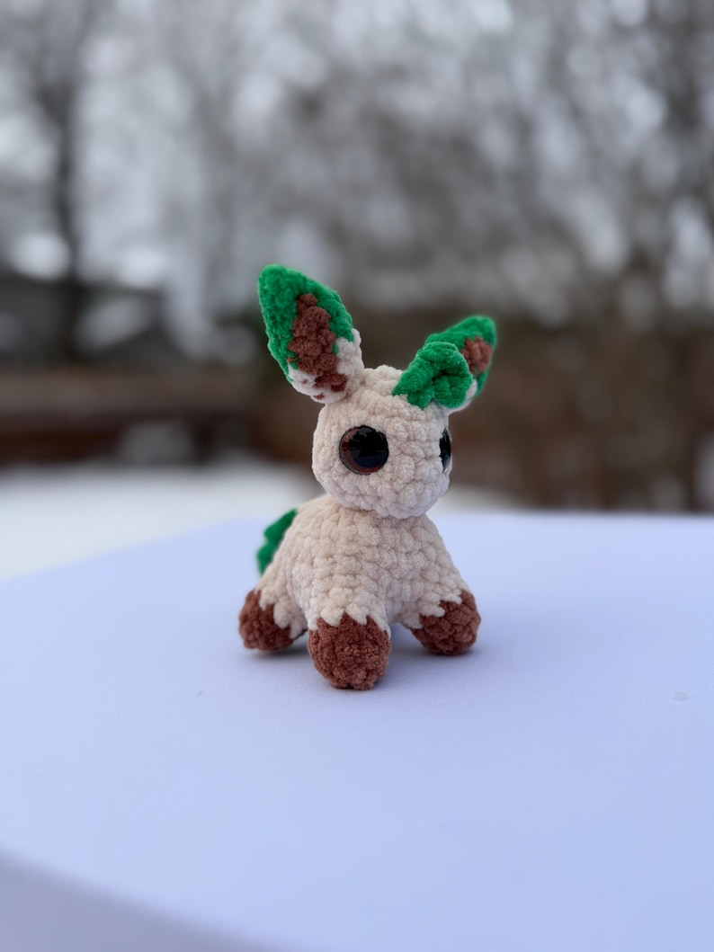 Puede incluir: Un peluche hecho a mano con ganchillo en forma de un peque&ntilde;o animal. El juguete tiene un cuerpo color crema, patas marrones y orejas verdes y marrones. Tiene ojos negros grandes y una peque&ntilde;a cola verde. El juguete est&aacute; sobre una superficie blanca.