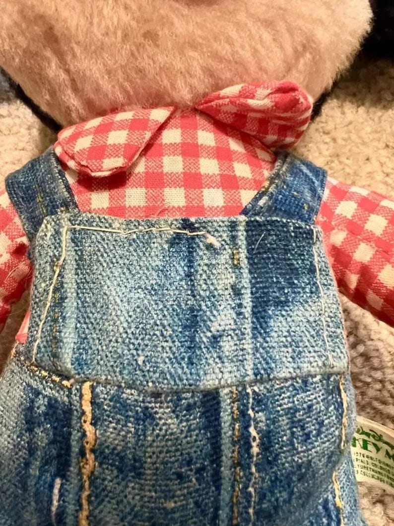 May include: A plush teddy bear wearing a pink and white checkered shirt and blue denim overalls. The bear has a brown fur coat and a stitched smile.