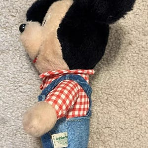 May include: A plush Mickey Mouse toy wearing blue denim overalls with a red and white checkered shirt. The toy has a green tag that reads "KiddieStuff".