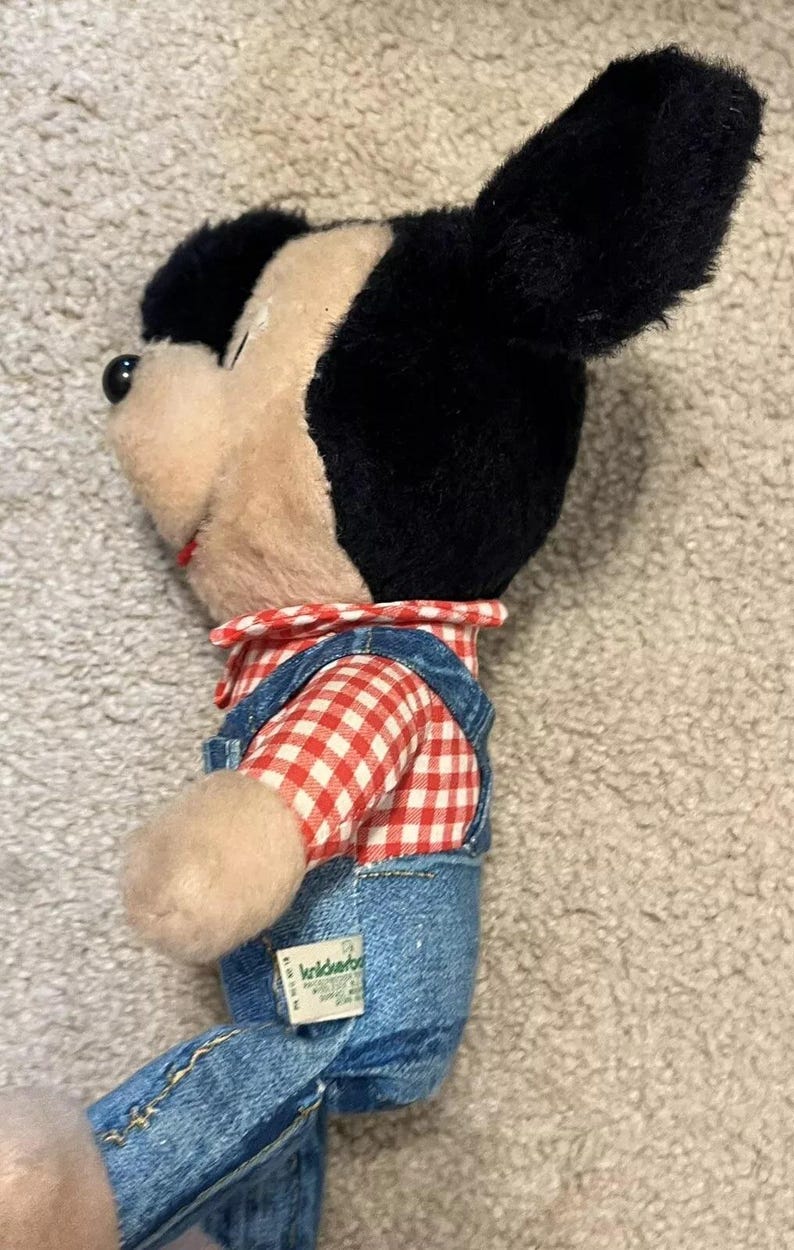 May include: A plush Mickey Mouse toy wearing blue denim overalls with a red and white checkered shirt. The toy has a green tag that reads "KiddieStuff".