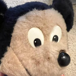May include: A vintage plush Mickey Mouse toy with black ears, brown fur, and white eyes. The toy is wearing a red and white checkered shirt and blue overalls.