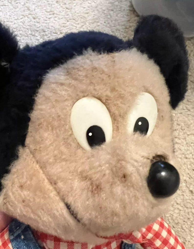 May include: A vintage plush Mickey Mouse toy with black ears, brown fur, and white eyes. The toy is wearing a red and white checkered shirt and blue overalls.
