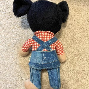May include: A black and white plush toy with large ears, wearing a red and white checkered shirt and blue denim overalls.