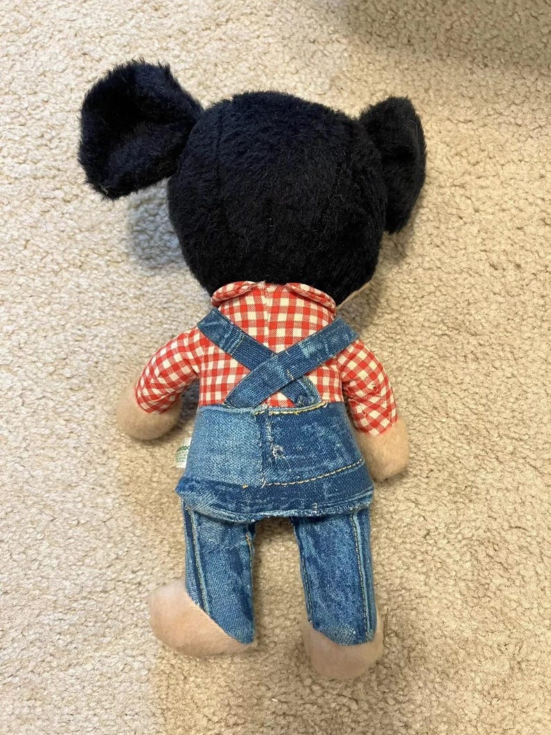 May include: A black and white plush toy with large ears, wearing a red and white checkered shirt and blue denim overalls.