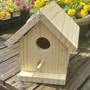 May include: A simple wooden birdhouse with a small entrance hole and a perch. The birdhouse is light brown and has a metal chain attached to the top.