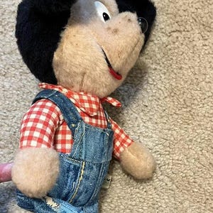 May include: A plush toy mouse wearing a red and white checkered shirt and blue denim overalls. The mouse has black ears and a brown face with a big smile.