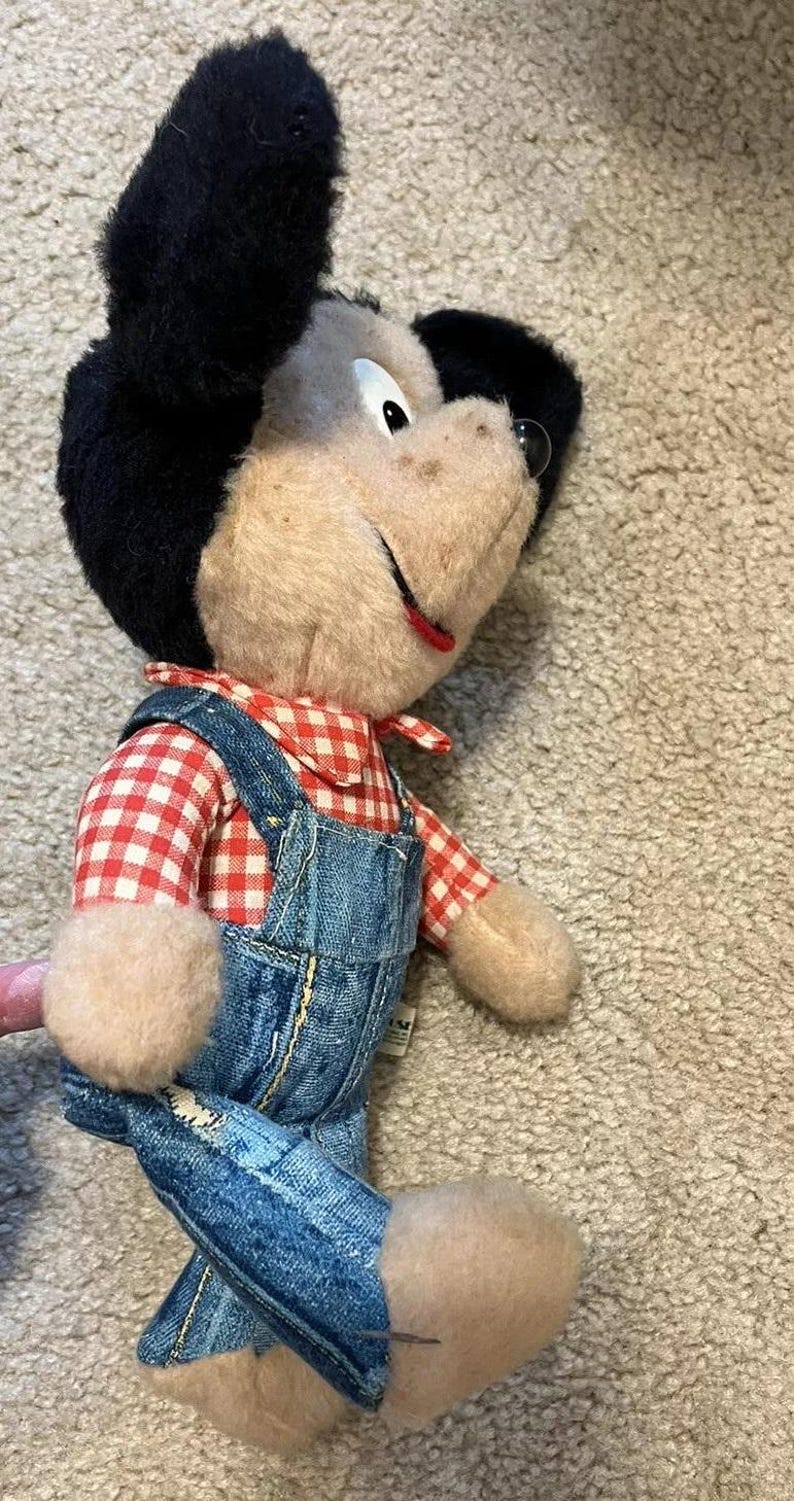 May include: A plush toy mouse wearing a red and white checkered shirt and blue denim overalls. The mouse has black ears and a brown face with a big smile.