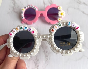 May include: Two pairs of flower-shaped sunglasses. One pair is white with black lenses and the text "BIG SIS" and "ELEANOR" on the frames. The other pair is pink with purple lenses and small flower accents.
