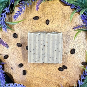 May include: A white bar of soap with brown coffee grounds embedded in it. The soap is on a wooden surface with coffee beans scattered around it.