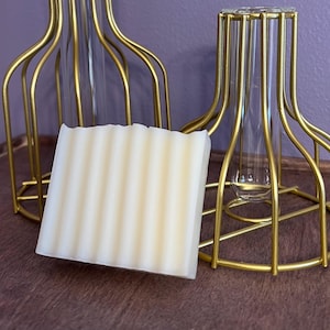 May include: A white bar of soap with a rippled texture, sitting on a wooden surface in front of two gold metal decorative stands.