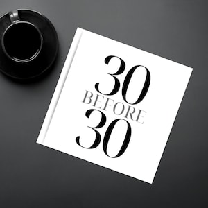 May include: A white book with the text "30 BEFORE 30" in black and gray. A black coffee cup and saucer are in the upper left corner, with a pen and keyboard nearby. The items are on a dark gray surface.