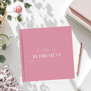 May include: A pink journal with the words "First Year of Retirement" in white script. A pink pen, a stack of books, a cup of coffee, and a decorative box are also on the white surface.
