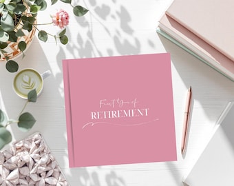 May include: A pink journal with the words "First Year of Retirement" in white script. A pink pen, a stack of books, a cup of coffee, and a decorative box are also on the white surface.