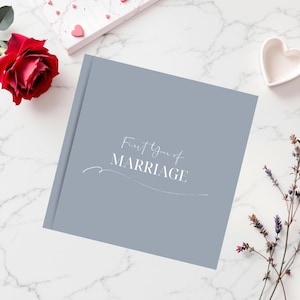 May include: A gray book with the text "First Year of Marriage" in white script. A red rose, a small white heart-shaped dish, and sprigs of purple flowers are arranged around the book on a white marble surface.