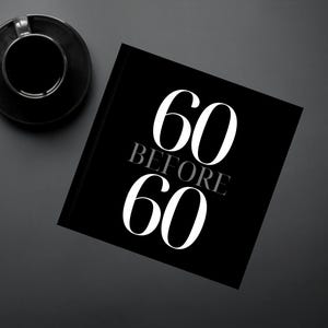 May include: A black notebook with the text "60 BEFORE 60" in white. A black coffee cup and saucer are in the upper left corner, and a pen is to the left of the notebook. The background is a dark grey surface.
