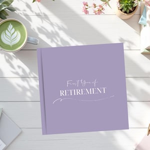 May include: A lavender-colored journal with the words "First Year of Retirement" in white script. The journal is on a white wooden surface, surrounded by a cup of matcha, a pen, and small plants.