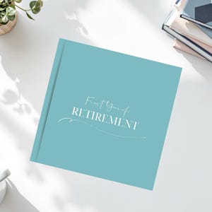 May include: A light blue book with the text "First Year of Retirement" in white script. The book is on a white surface with a small potted plant and a cup of coffee in the background. Other books are stacked nearby.