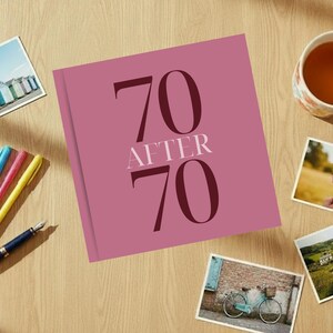 May include: A mauve book titled "70 After 70" is surrounded by colorful pens, a fountain pen, postcards, and a floral teacup on a wooden surface. The postcards depict beach huts, ice cream, and a bicycle.