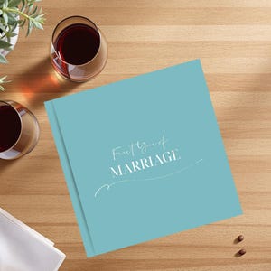 Puede incluir: Un libro de color turquesa con la inscripci&oacute;n "First Year of MARRIAGE" en escritura blanca. Dos copas de vino tinto y una peque&ntilde;a planta en maceta est&aacute;n sobre una superficie de madera.