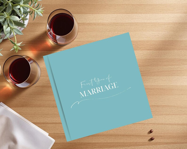 Puede incluir: Un libro de color turquesa con la inscripci&oacute;n "First Year of MARRIAGE" en escritura blanca. Dos copas de vino tinto y una peque&ntilde;a planta en maceta est&aacute;n sobre una superficie de madera.