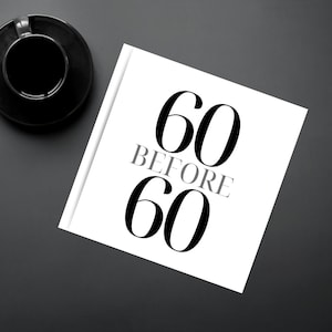 May include: A white book with the text "60 BEFORE 60" in black and grey. A black coffee cup and saucer are in the upper left corner, with a pen and keyboard nearby. The items are on a dark grey surface.