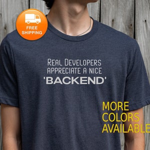 Op de afbeelding: Een donkergrijs T-shirt met de tekst "Real Developers Appreciate a Nice 'BACKEND'" in wit gedrukt. De tekst "MORE COLORS AVAILABLE" is in geel gedrukt aan de onderkant van het shirt.