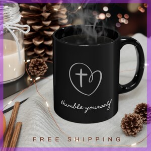 May include: Black ceramic coffee mug with a white heart and cross design. The mug has the text "Humble yourself" written on it.