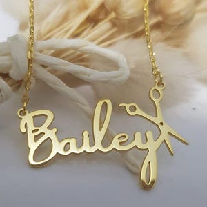 May include: Gold necklace featuring the name "Bailey" in a cursive script with a pair of scissors incorporated into the design.
