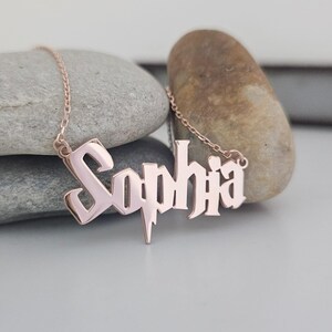 May include: Rose gold necklace with the name "Sophia" in a decorative font. The pendant is suspended from a fine chain. The necklace is shown against a light grey backdrop with two stones.