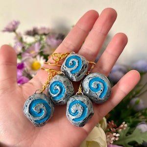 Puede incluir: Seis llaveros grises con forma de piedra con un diseño de espiral azul. Cada llavero tiene una cadena dorada. Los llaveros se sostienen en una mano, con un fondo floral borroso.