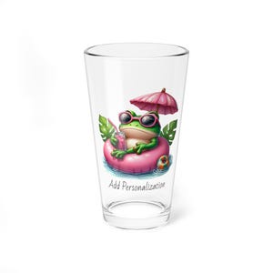 May include: A clear glass with a green frog wearing sunglasses and a pink inflatable ring, holding a pink drink. The frog is under a pink umbrella and the glass has the text "Add Personalization".