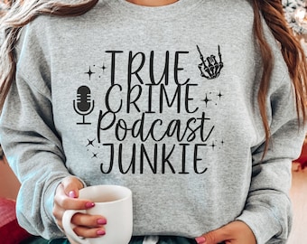 May include: A gray sweatshirt with the text "TRUE CRIME PODCAST JUNKIE" in black lettering. The text is surrounded by small stars and a skeleton hand making a rock and roll hand gesture. A microphone icon is also included in the design.