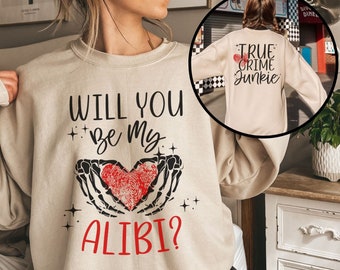 May include: A beige sweatshirt with the text "Will you be my alibi?" printed on the front. The text is in black with a red heart and black skeletal hands holding the heart. The back of the sweatshirt has the text "True Crime Junkie" printed in black with a red heart.
