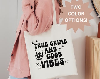 May include: Cream-colored tote bag with the text "TRUE CRIME AND GOOD VIBES" in black. The design includes a skeleton hand giving the middle finger. The bag has long, cream-colored straps.