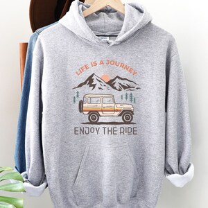 May include: Grey hooded sweatshirt with a graphic of a vintage jeep driving through a mountain landscape. The text "Life is a Journey, Enjoy the Ride" is printed on the sweatshirt.